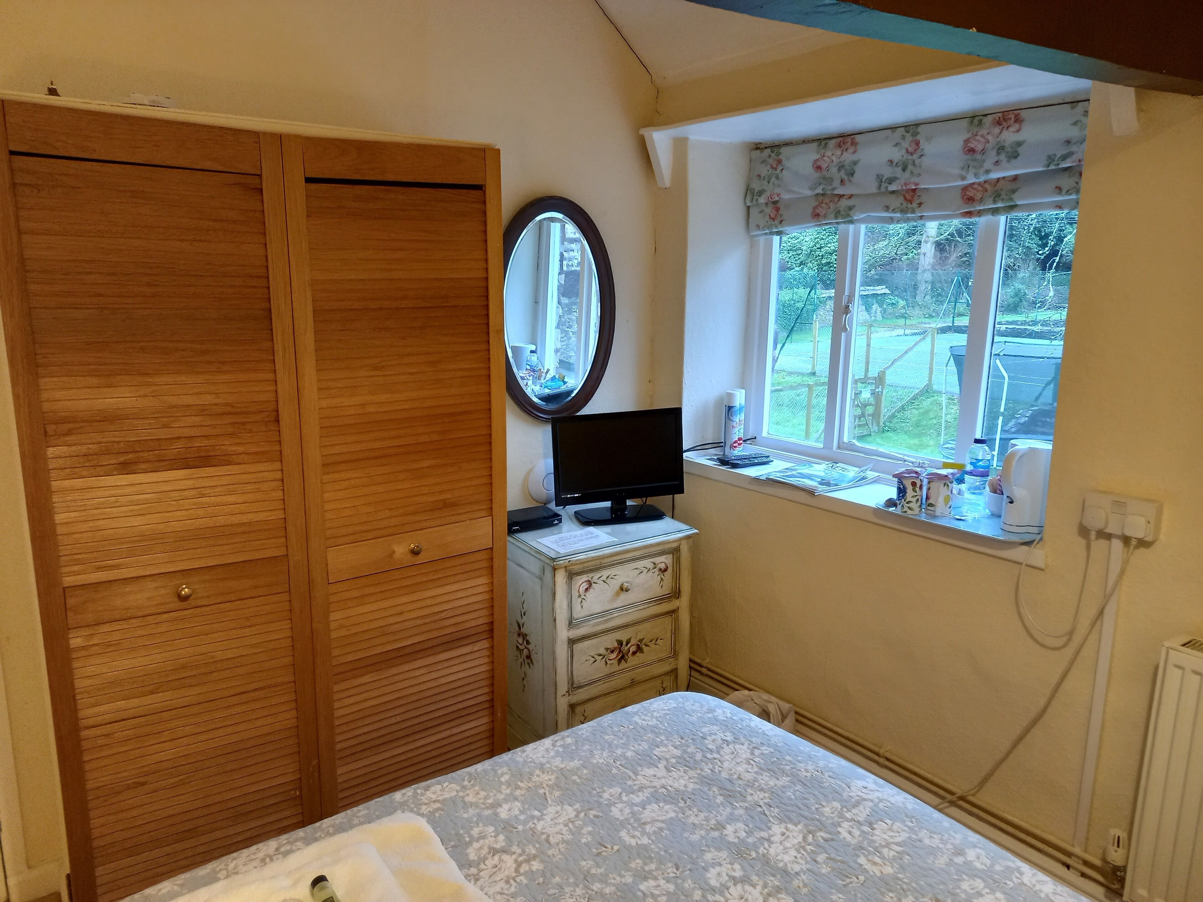 A bedroom with a wooden wardrobe, a bedside table with a TV, a window with a floral valance, and a radiator.