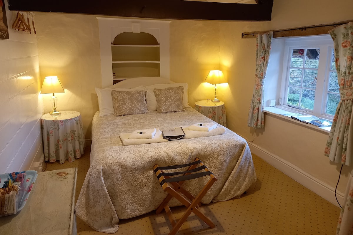 A neatly made bed with floral-patterned bedding, two bedside lamps, a small table with a lamp, a window with floral curtains, and a wooden chair.