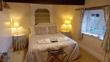 Una cama bien hecha con ropa de cama de estampado floral, dos lámparas de noche, una mesita con lámpara, una ventana con cortinas florales y una silla de madera.