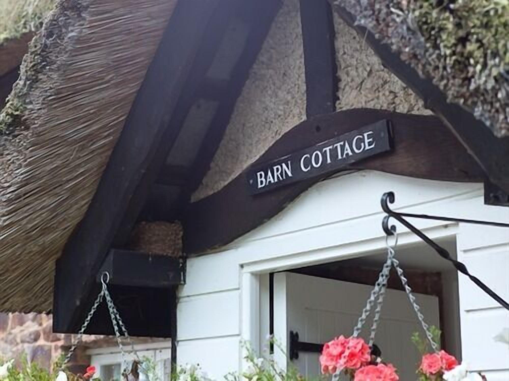 A rustic building with a 'Barn Cottage' sign, a thatched roof, and hanging flower baskets.