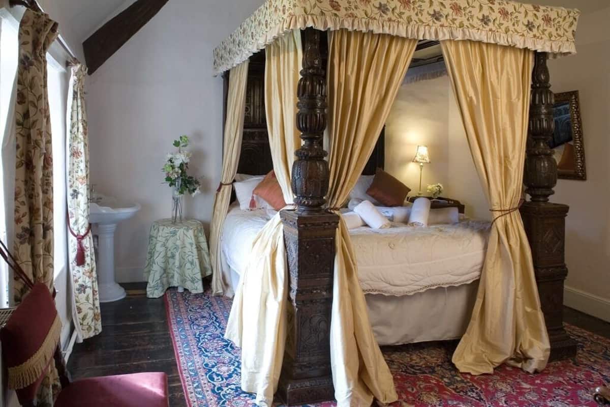 A traditional four-poster canopy bed with a floral bedspread, a bedside lamp, and a small table with a vase of flowers.