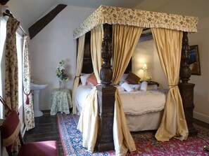 A traditional four-poster canopy bed with a floral bedspread, a bedside lamp, and a small table with a vase of flowers. - Bickleigh Castle Hotel (Tiverton)