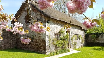 A stone building with a thatched roof, surrounded by lush greenery and blooming pink flowers.