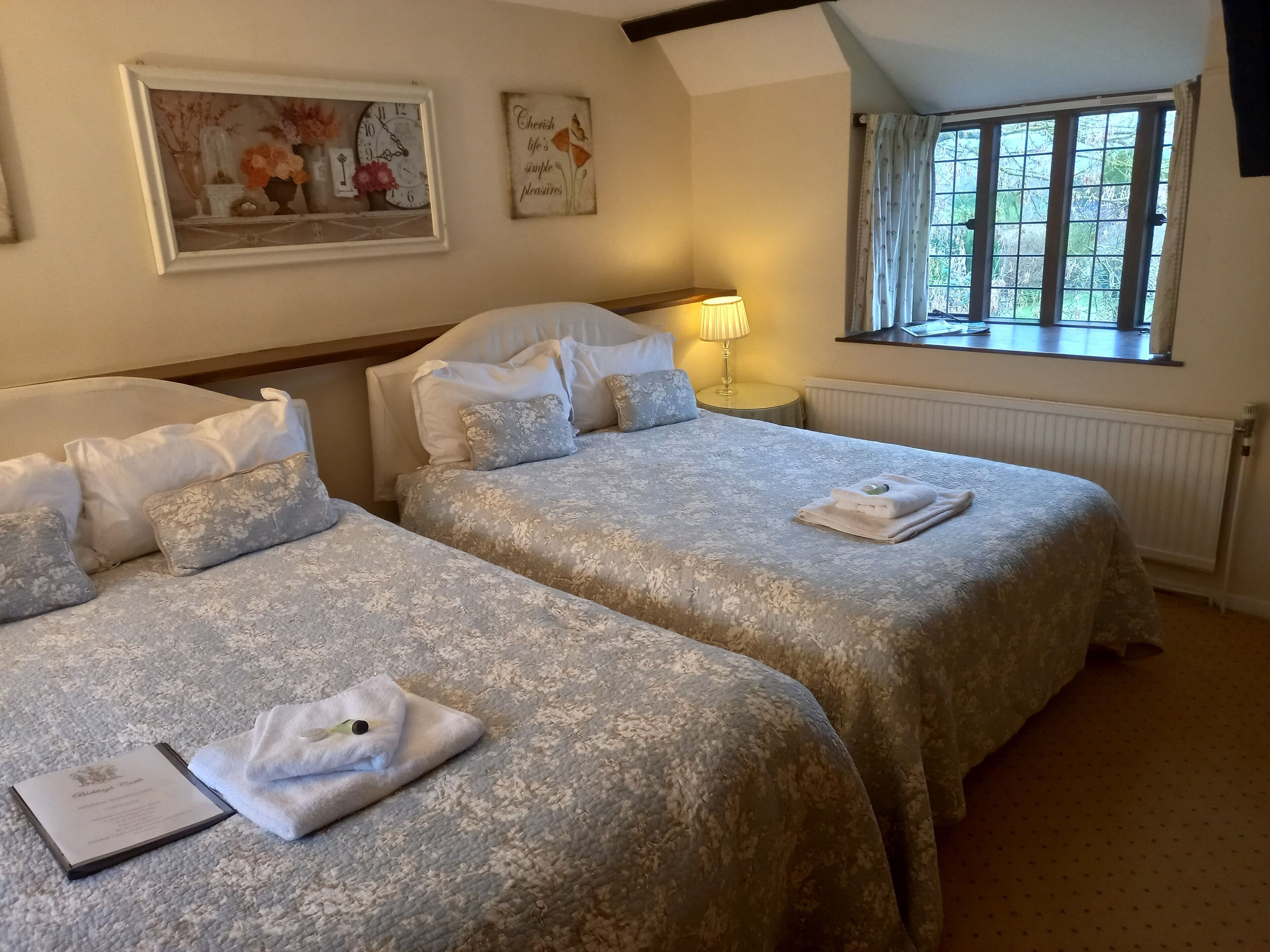 A hotel room with two beds, a framed floral painting, a clock, a bedside lamp, and a window with curtains.