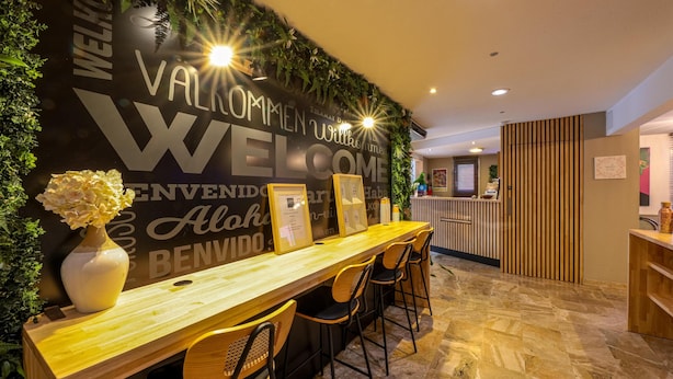 A reception area with a wooden reception desk, several chairs, a wall with a welcome sign, and a potted plant. - Ostal Pau Université - Sure Hotel Collection by Best Western (Pau)