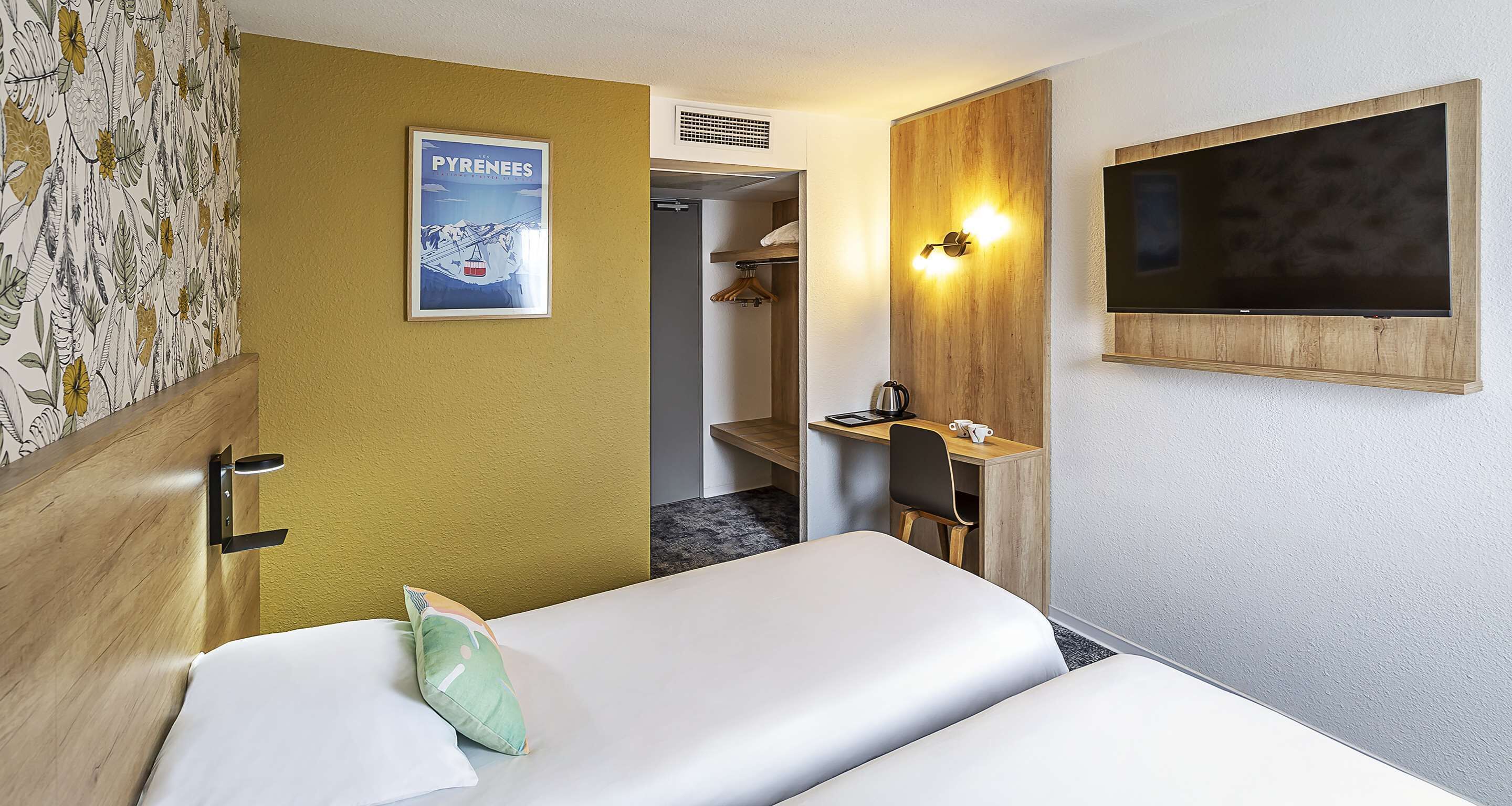 A hotel room with two beds, a wooden desk with a chair, a mounted TV, and a poster on the wall.