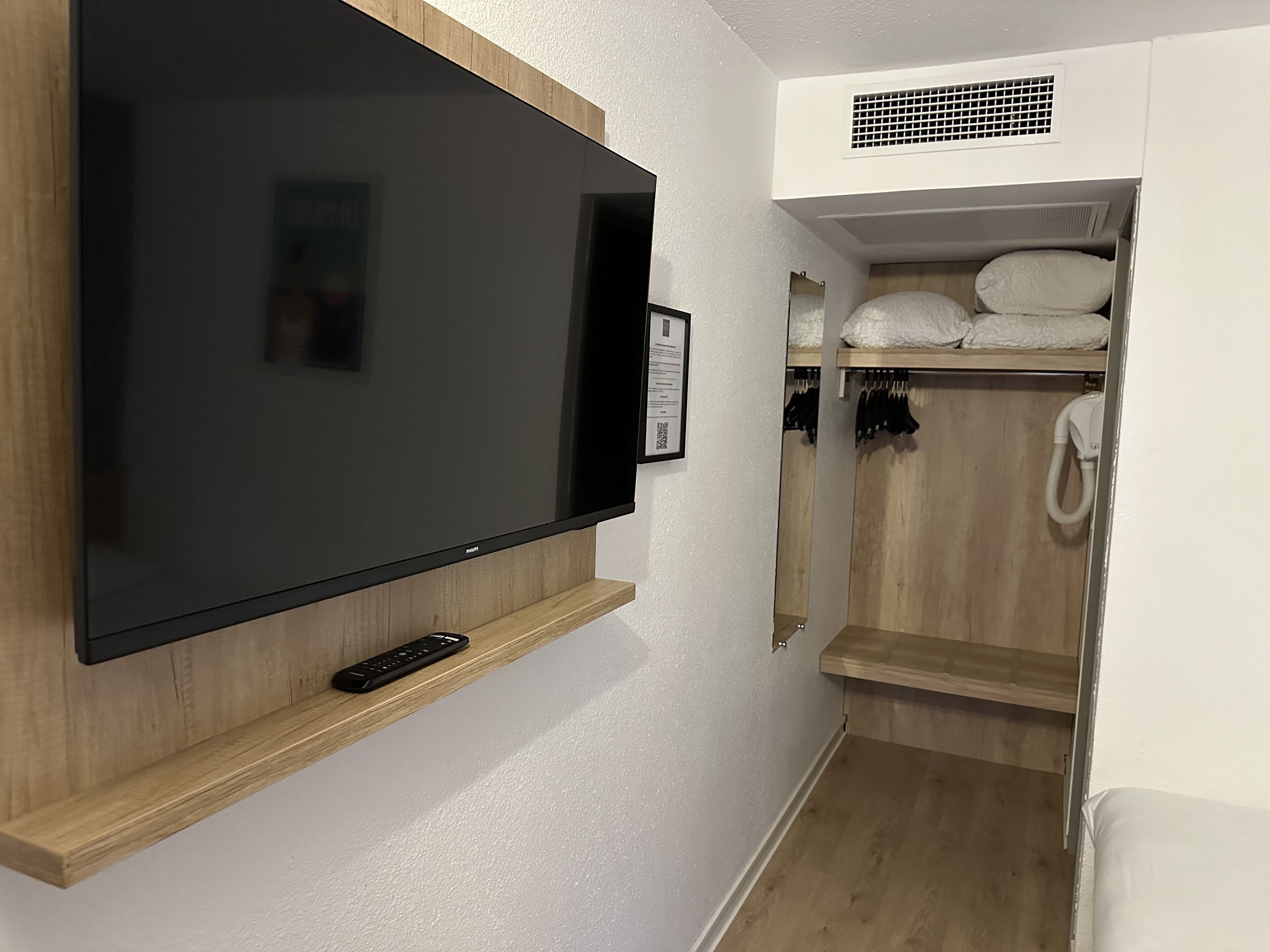Una habitación moderna con un televisor de pantalla plana grande montado en un estante de madera, un control remoto pequeño, una pared blanca, un armario de madera con ropa colgada y una rejilla blanca en el techo.