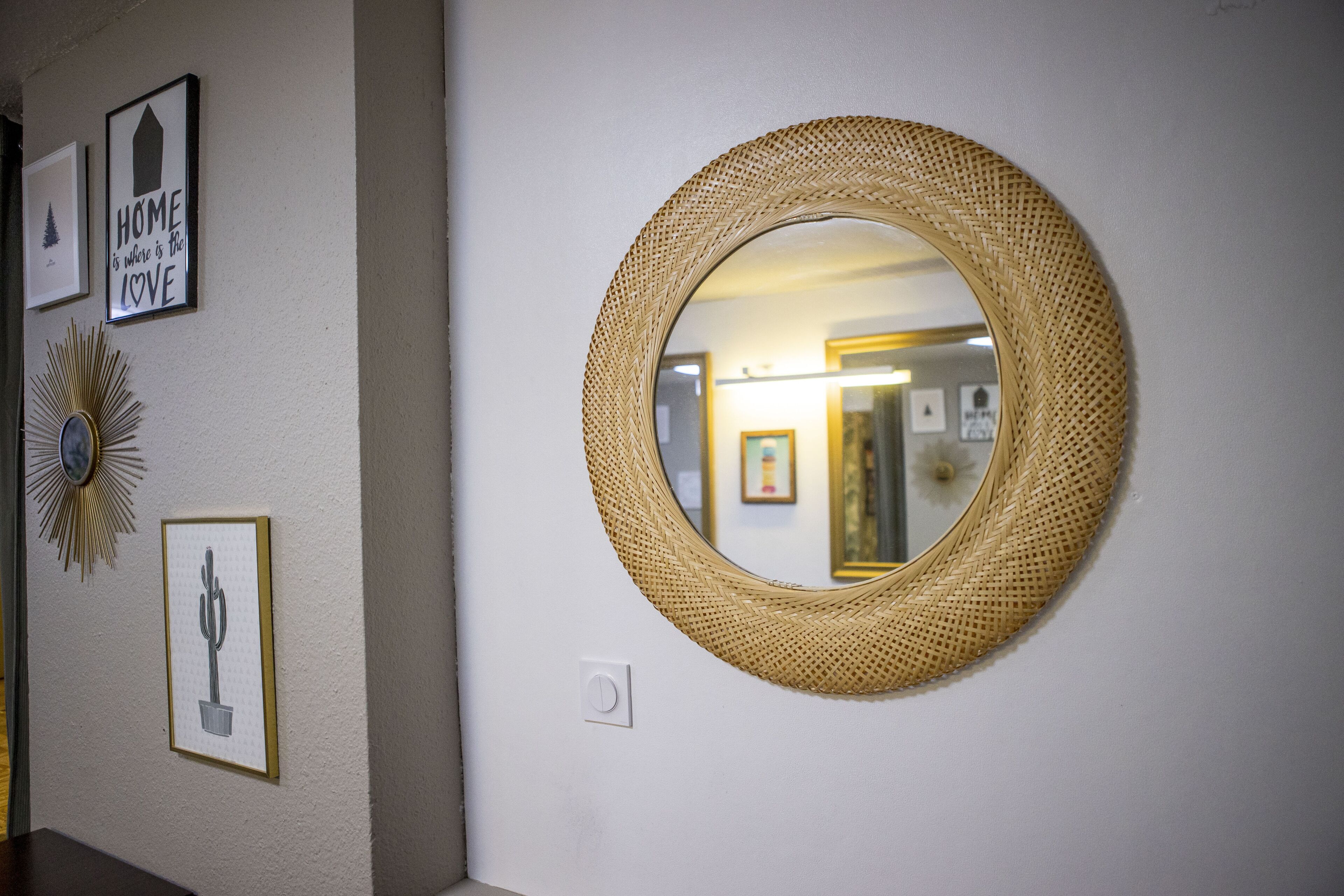 A wall with a round woven mirror, framed pictures, and a wall sconce.