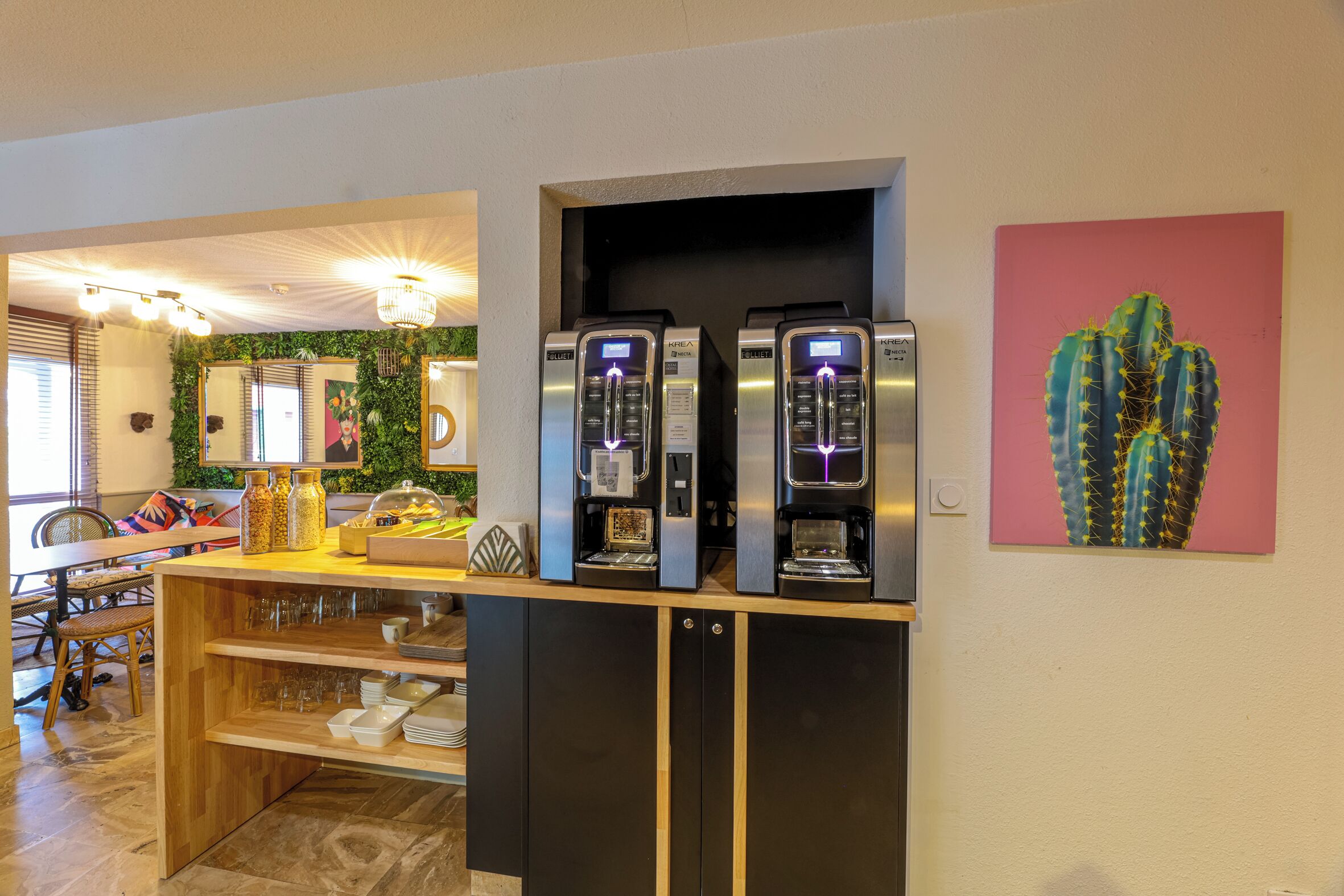 Un coin cuisine avec un espace dédié au café, comprenant deux machines à café, une œuvre d’art représentant un cactus accrochée au mur et un comptoir en bois avec de la verrerie.