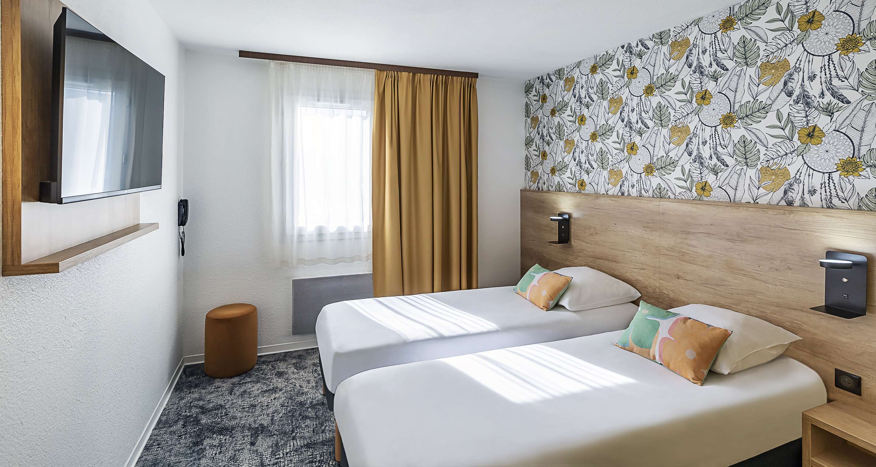 A hotel room with two beds, a TV, a wooden headboard, a patterned wall, and a window with curtains.
