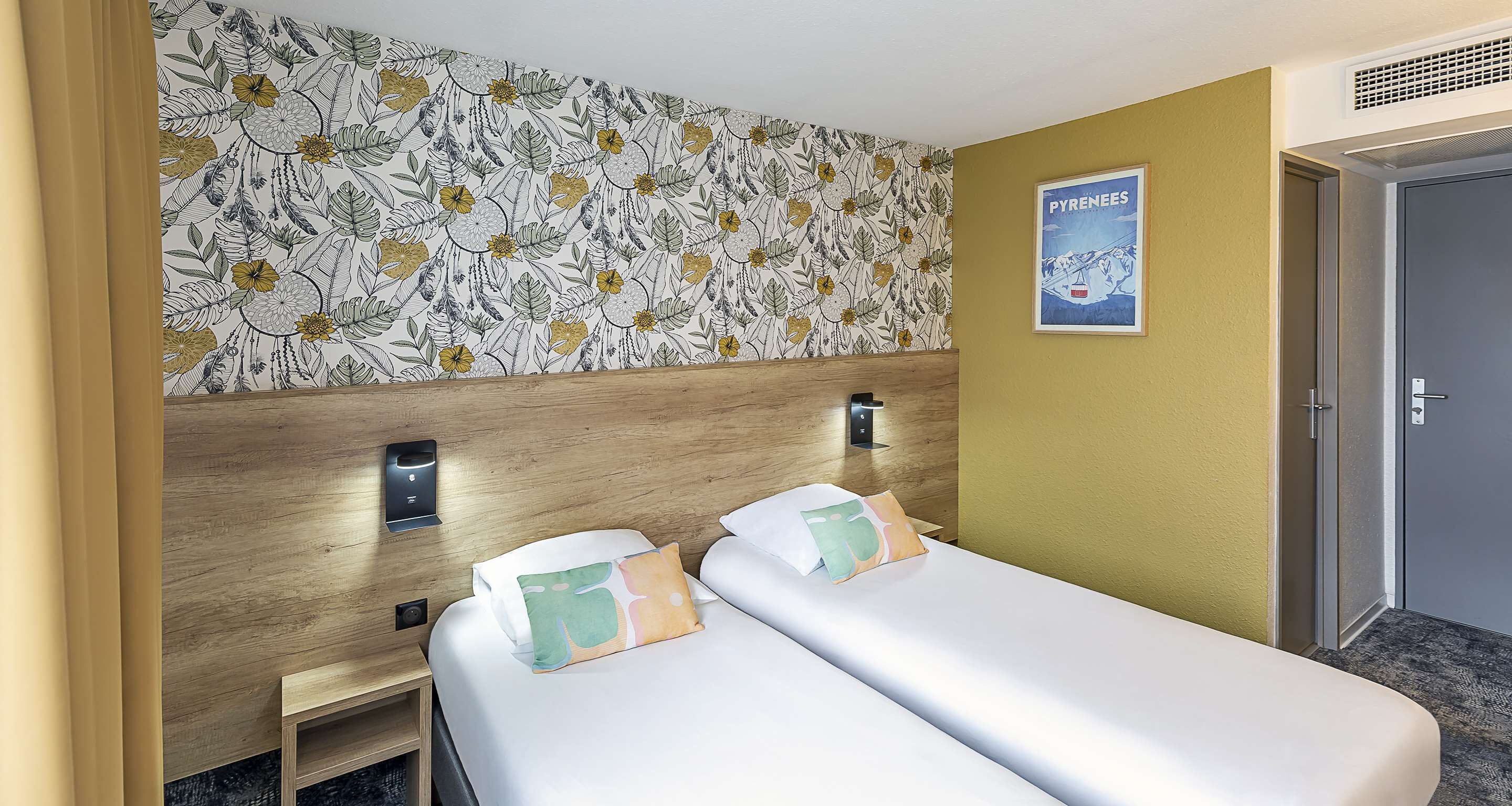 Et hotelværelse med to senge, en træsenggavl, blomstret tapet, et billede på væggen og en dør.