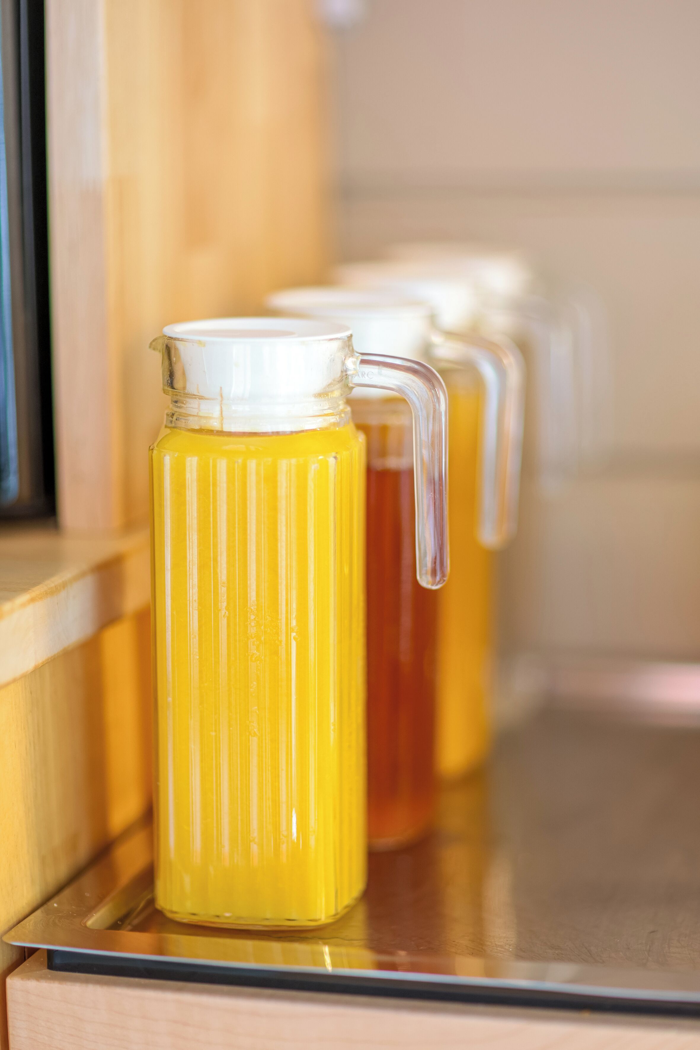 A stainless steel kitchen counter with a glass pitcher filled with a yellow liquid, likely juice or tea, and a matching glass pitcher in the background.