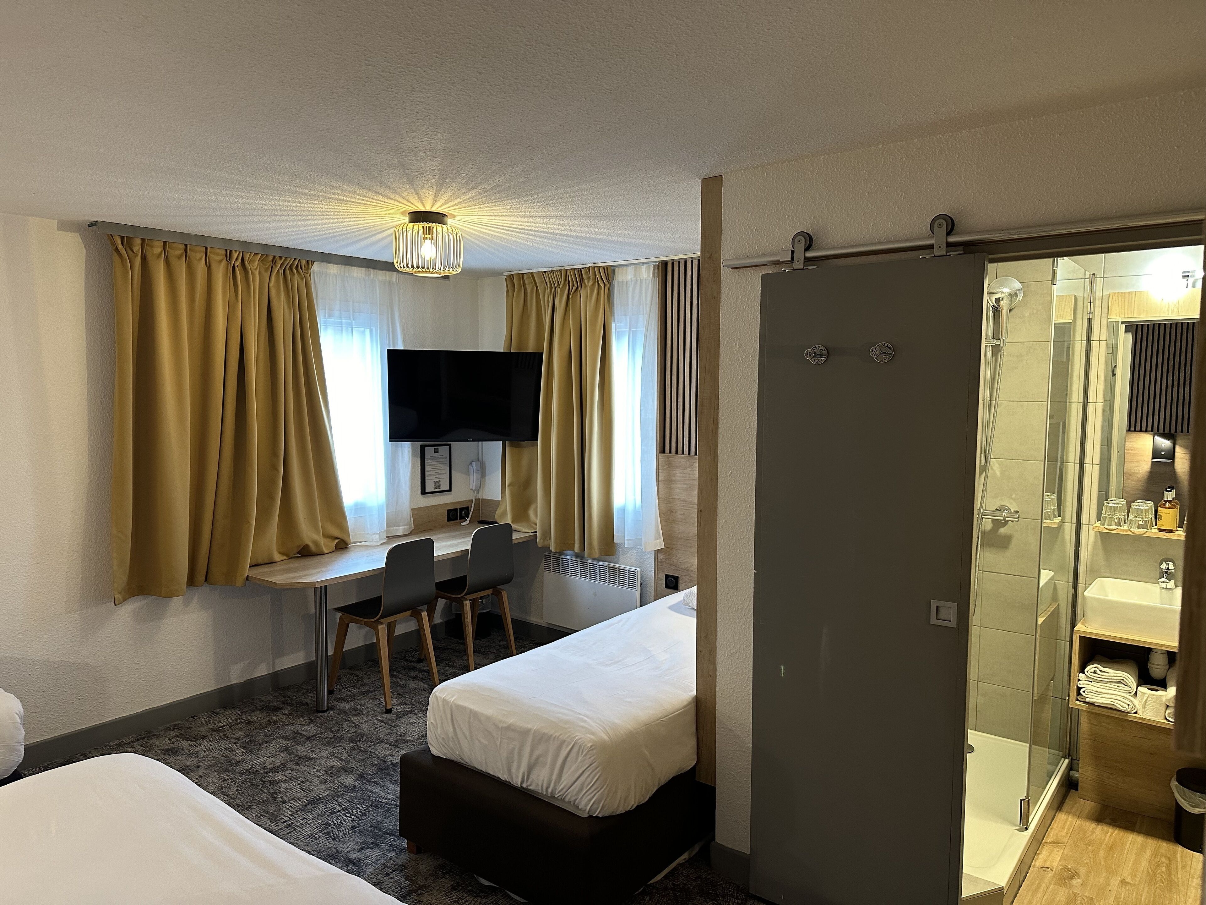 Quarto de hotel com cama, escrivaninha com cadeira, televisão e área de chuveiro.