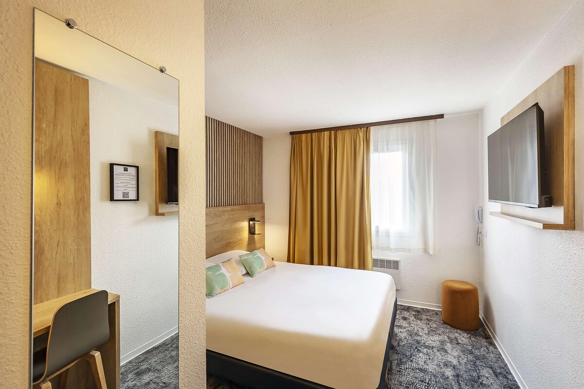 Hotellihuone, jossa on sänky, työpöytä, tuoli, televisio, ikkuna verhoilla ja pieni jakkara.