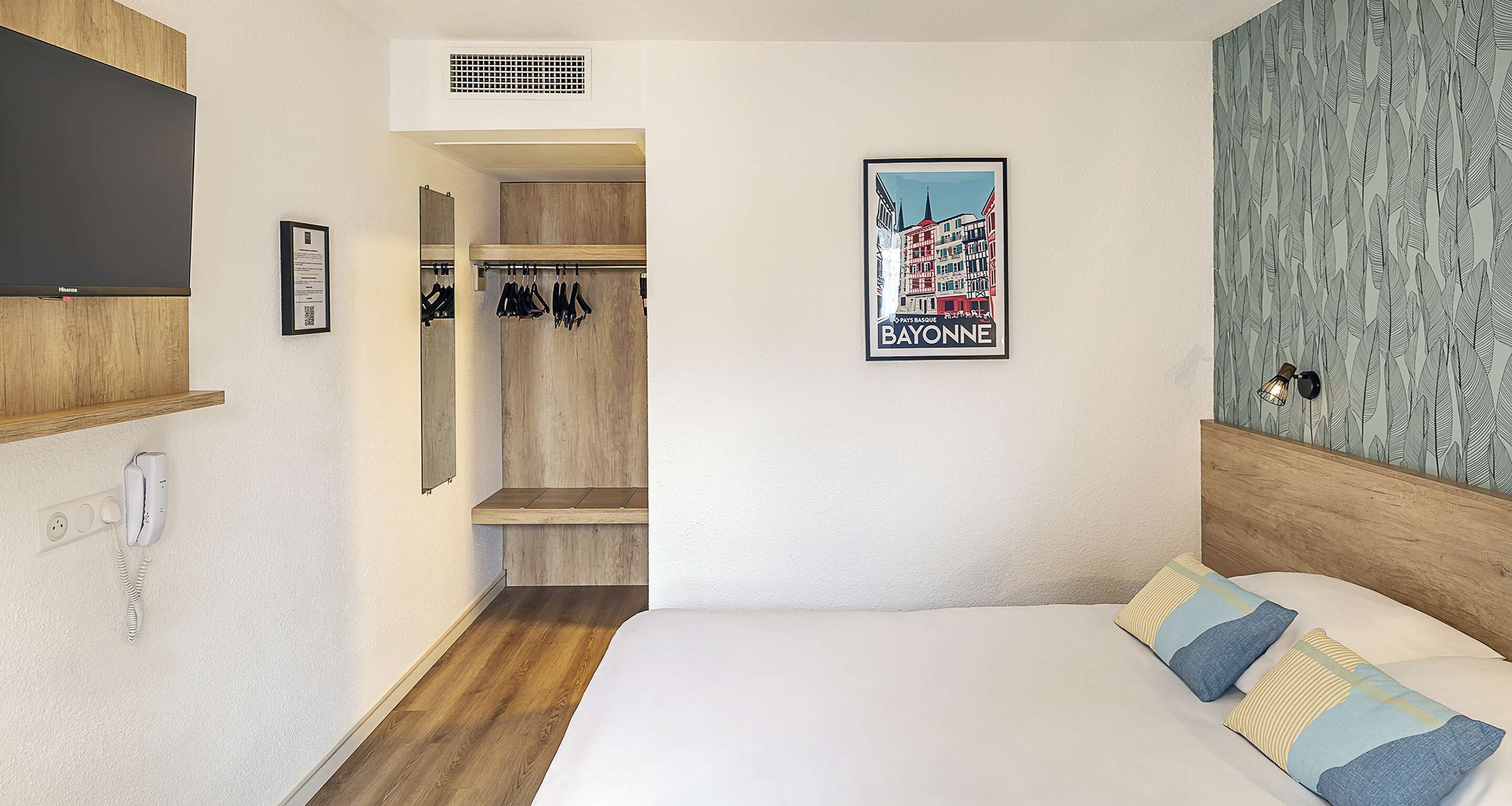 A hotel room with a bed, a wardrobe, a mirror, a framed picture of Bayonne, and a telephone on the wall.
