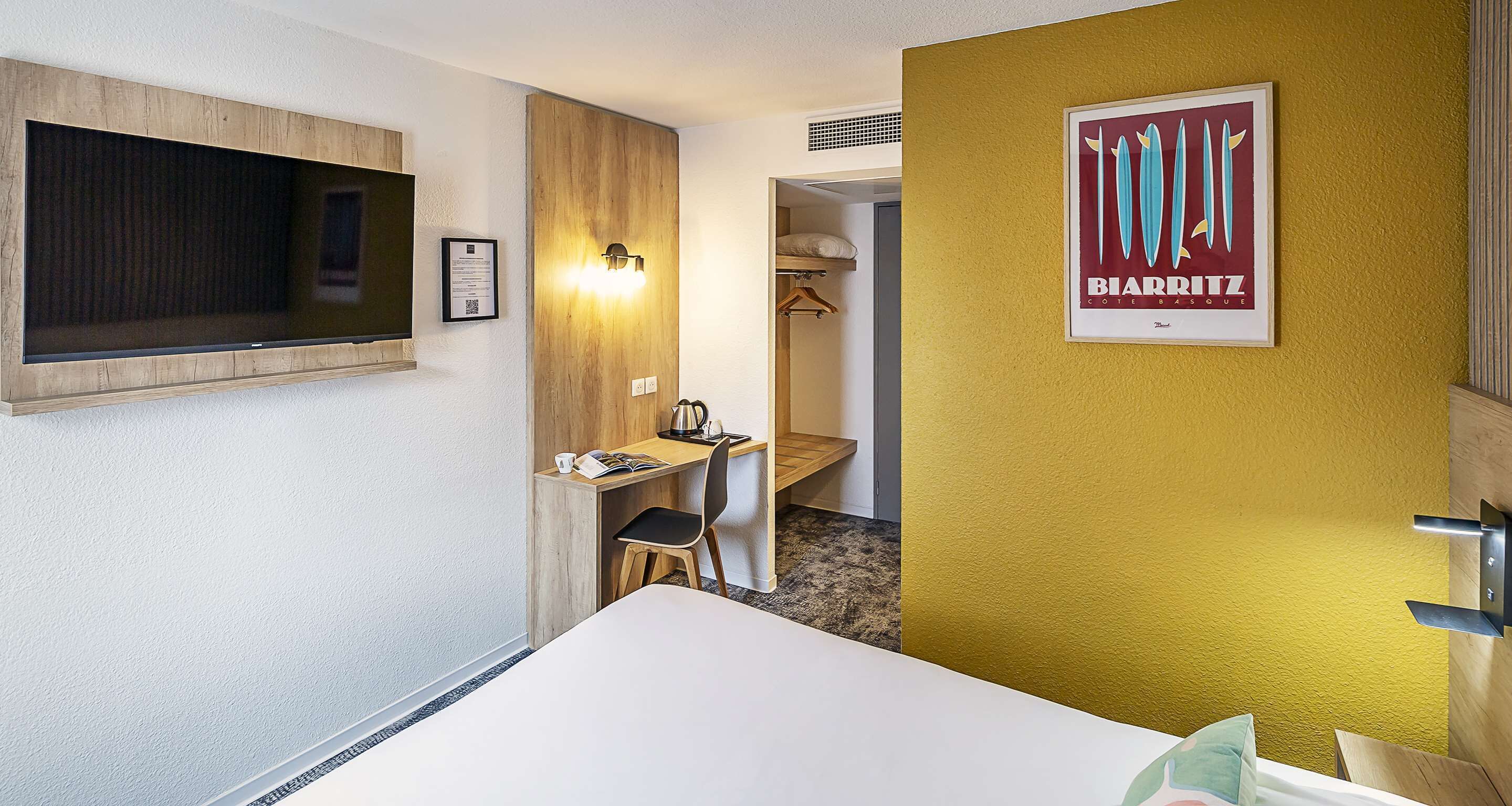Une chambre d’hôtel avec un lit, un bureau et une chaise, un téléviseur fixé au mur et une affiche sur la porte.