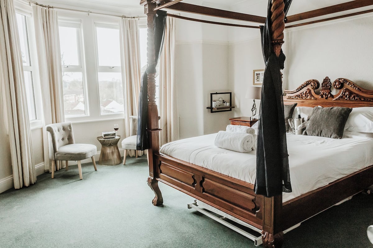A four-poster canopy bed with a wooden headboard, a bedside table with a bottle and glasses, and a chair by the window.
