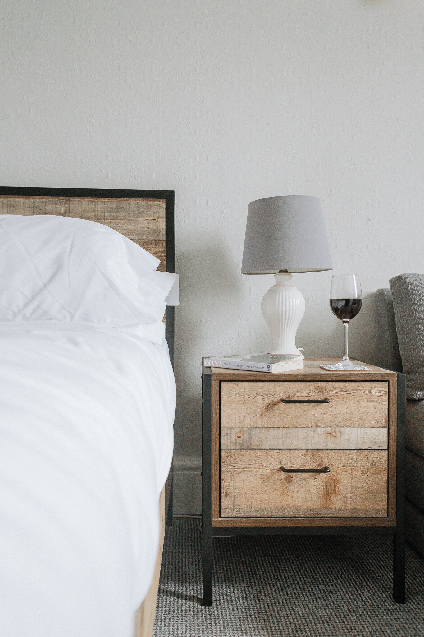 Una cama bien hecha con sábanas blancas, una mesita de noche blanca con una lámpara y una copa de vino tinto, y una silla gris en una habitación sencilla con cabecera de madera y paredes lisas.