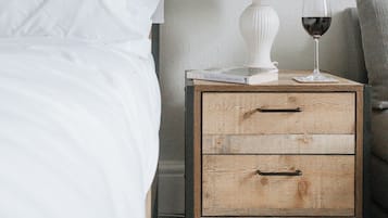 Una cama bien hecha con sábanas blancas, una mesita de noche blanca con una lámpara y una copa de vino tinto, y una silla gris en una habitación sencilla con cabecera de madera y paredes lisas.