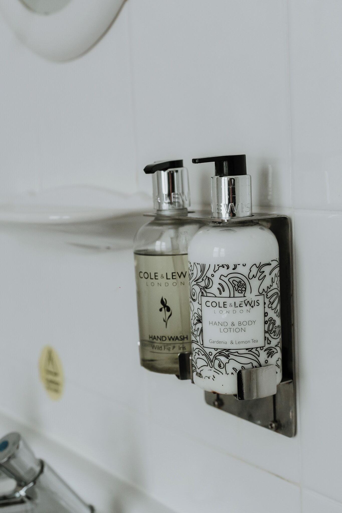 Two bottles of Cole & Lewis hand and body lotion mounted on a white tiled wall.