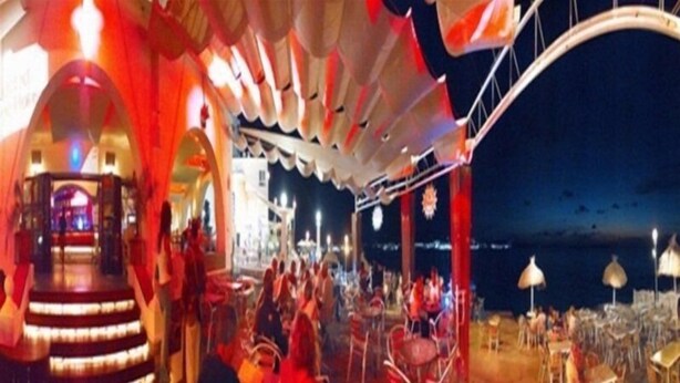 Un espace de restauration en plein air, aménagé pour le soir, avec des tables et des chaises, une scène dotée d’une plateforme circulaire et une grande structure ressemblant à une tente qui le surplombe.
