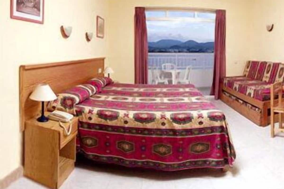 Quarto de hotel com cama, mesa de cabeceira, uma cadeira e vista para as montanhas pela janela.