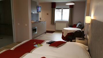 A hotel room with a bed, a desk, a chair, a TV, a window with curtains, a radiator, and a kitchenette.