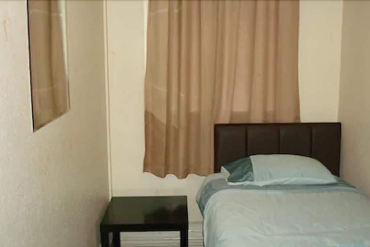 Quarto pequeno com uma cama de solteiro, cabeceira escura, mesa de cabeceira e janela com cortinas.