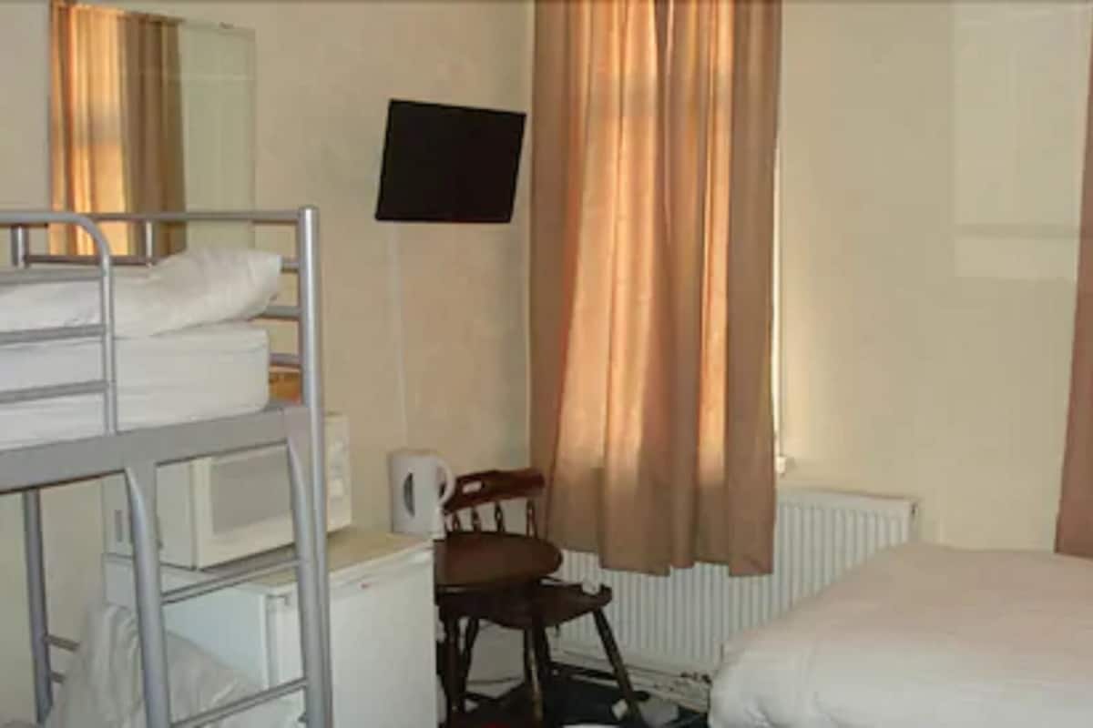 Quarto de hotel com beliches, uma escrivaninha, uma cadeira, um radiador e uma TV na parede.
