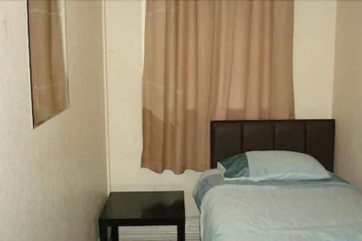 Quarto pequeno com cama de solteiro, cabeceira escura, mesa de cabeceira e janela com cortinas bege.
