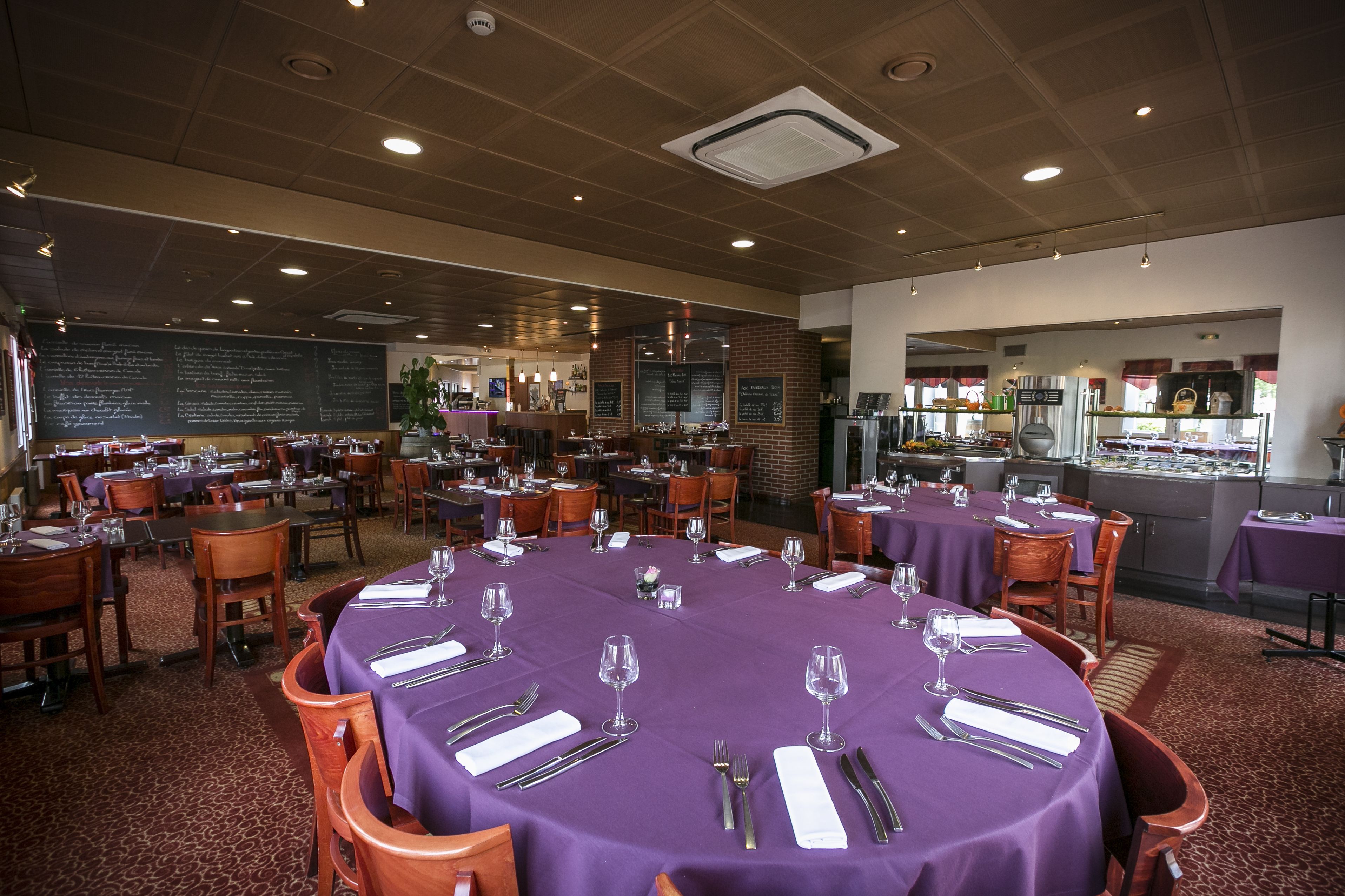 A restaurant with multiple tables set for dining, a kitchen area visible in the background, and a chalkboard menu on the wall.