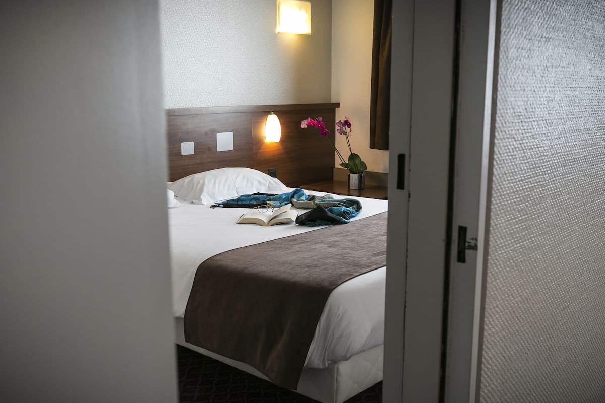A hotel room with a bed, a nightstand, a wall-mounted lamp, and a window with curtains.