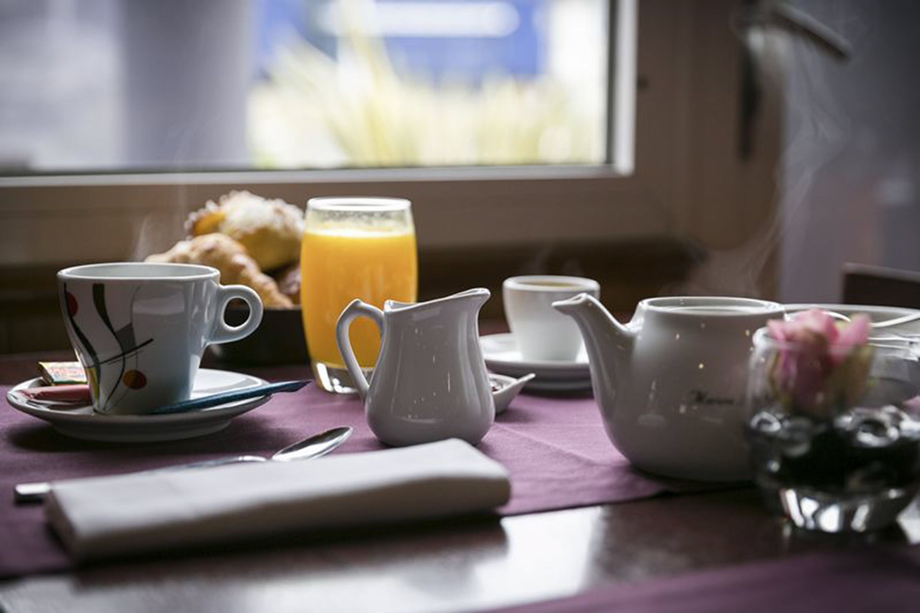 Une table près d’une fenêtre est dressée pour le petit-déjeuner, avec une tasse de café, un pichet, une théière et un muffin.