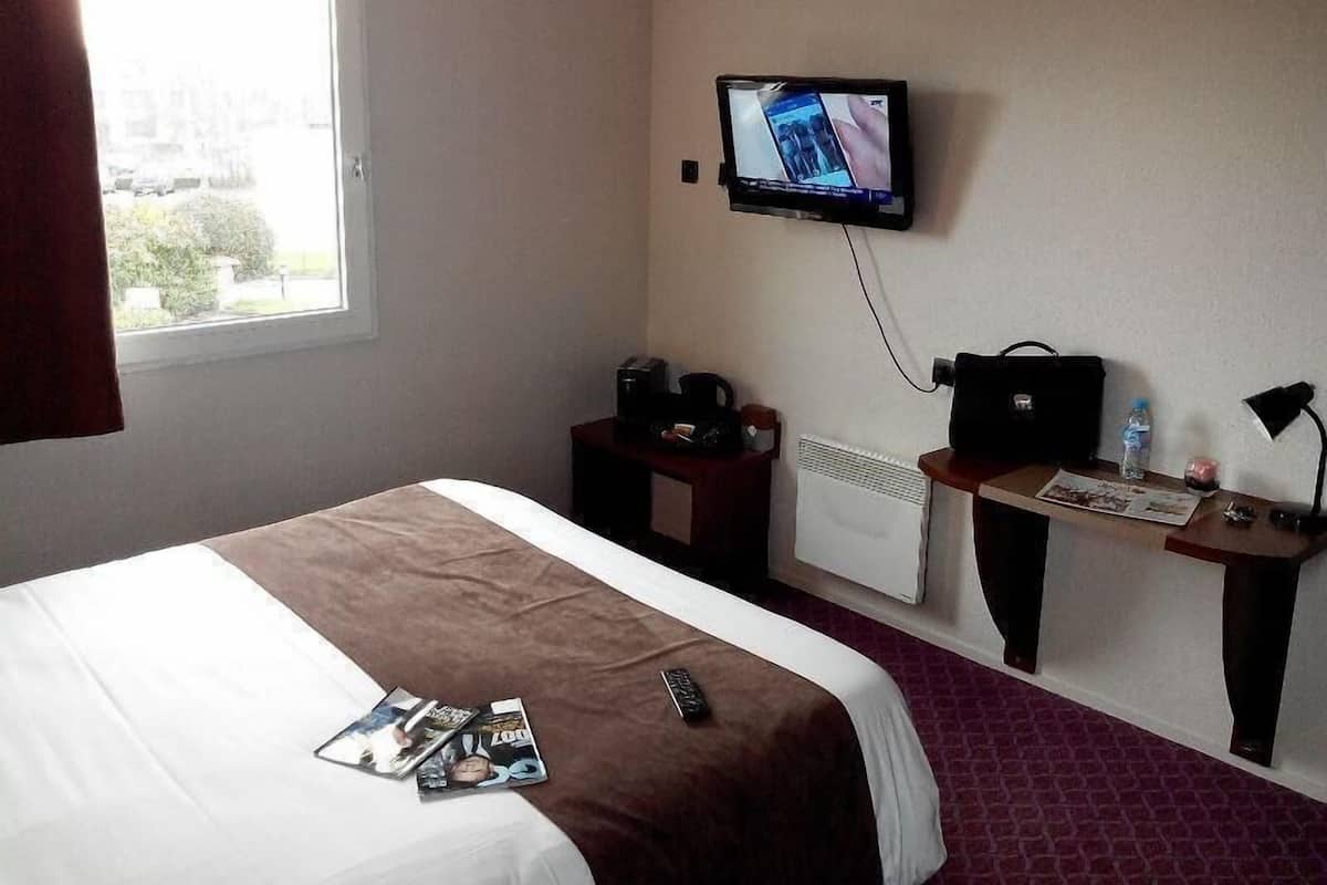 A hotel room with a bed, a TV mounted on the wall, a desk with a chair, a radiator, and a window with curtains.
