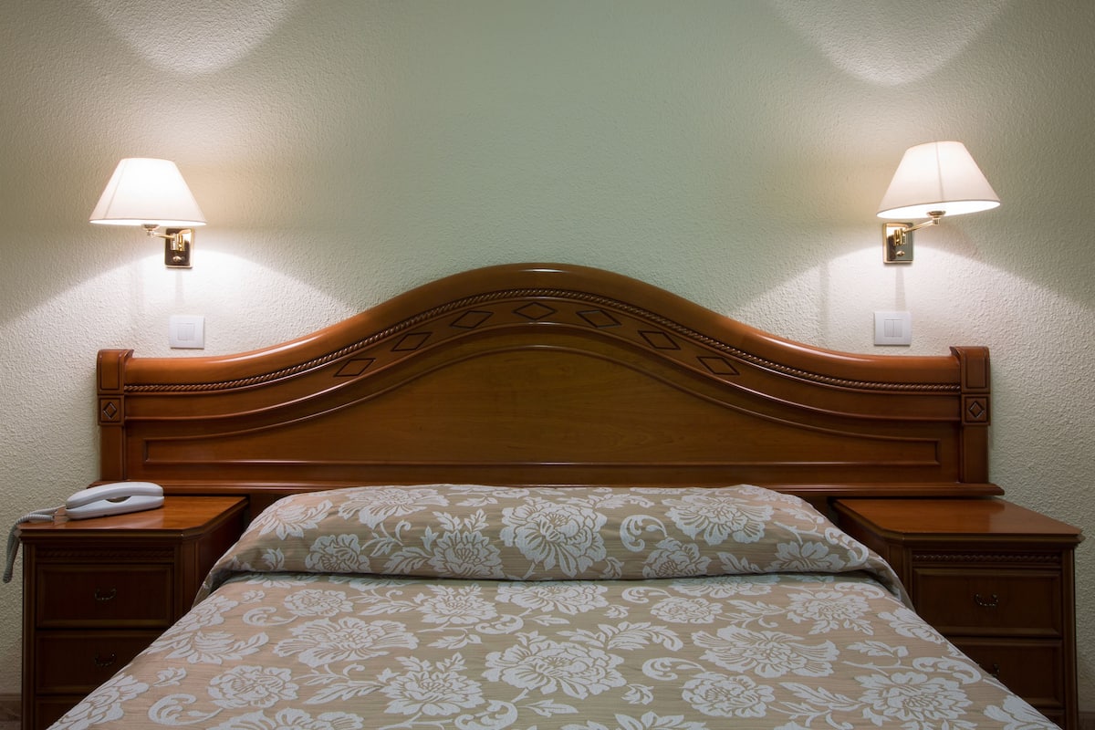 Una cama bien hecha con una colcha de estampado floral, flanqueada por dos mesitas de noche con lámparas, y un cabecero de madera con un diseño decorativo.