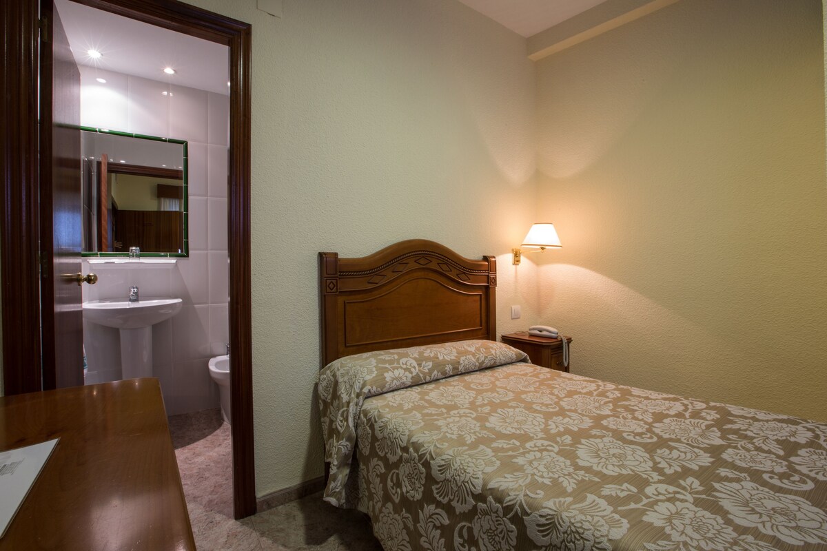 A hotel room with a wooden bed, a bedside lamp, a small bedside table, a mirror, a sink, and a toilet.