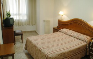 A bedroom with a bed, a bedside table with a lamp, a wooden nightstand, a chair, a desk with a television, and a radiator. - Hotel Riviera (Córdoba)