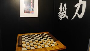 A chessboard on a wooden table against a black wall with a framed picture and Chinese characters. - Street Art Hotel (Rouen)