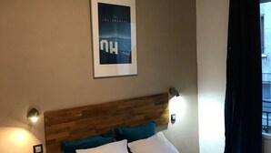 A hotel room with a wooden headboard, a bed with white and teal pillows, a small wooden table, and a poster on the wall. - Street Art Hotel (Rouen)
