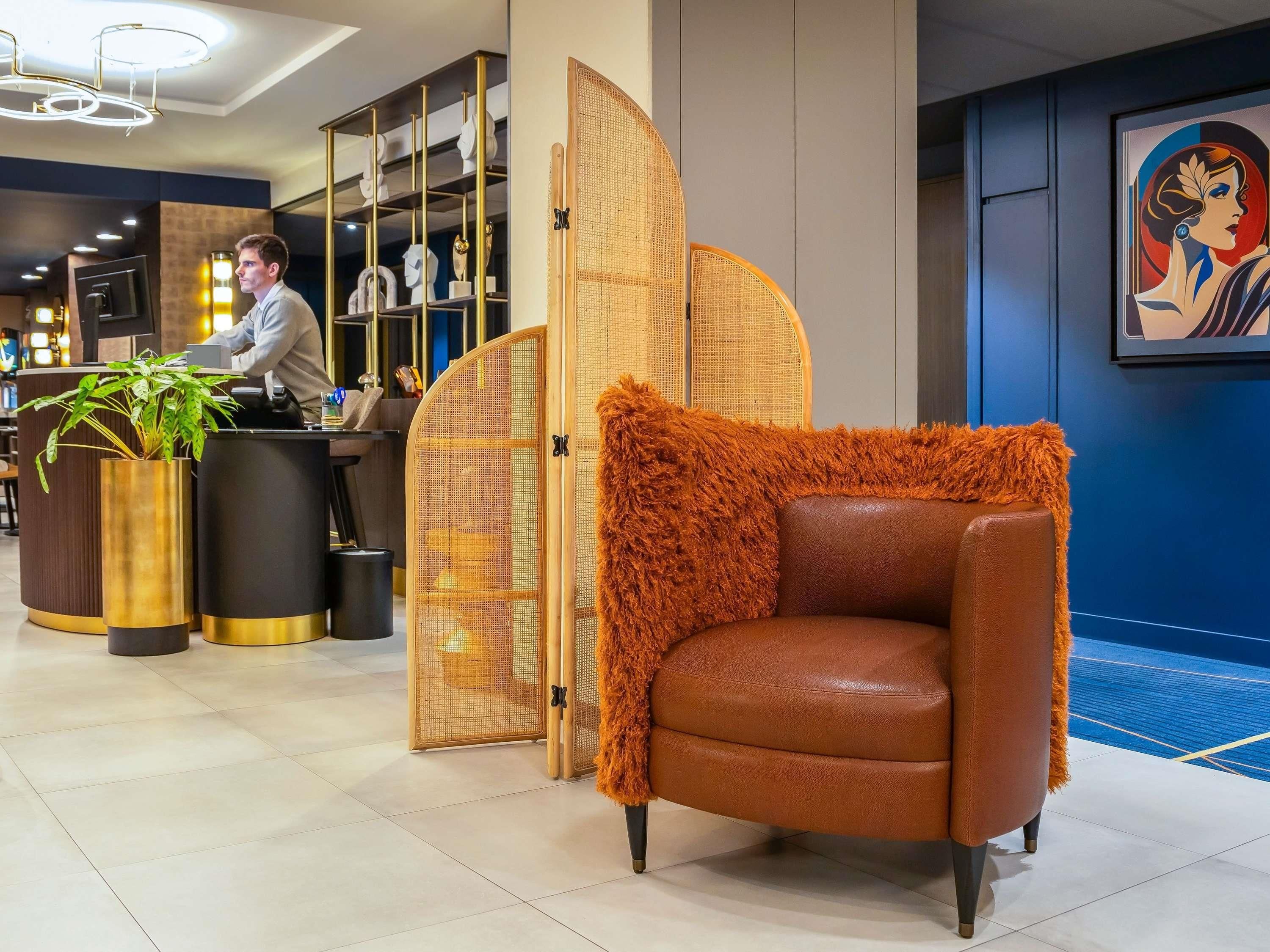 Een receptiezone met een balie, een plant, een decoratieve stoel en een scheidingswand met een geweven patroon.