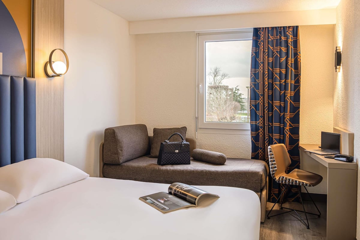 A hotel room with a bed, a desk with a laptop, a chair, a window with curtains, and a wall-mounted lamp.