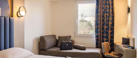 A hotel room with a bed, a desk with a laptop, a chair, a window with curtains, and a wall-mounted lamp.
