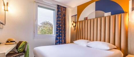Habitación de hotel con cama, escritorio, silla, ventana y pared con diseño geométrico.
