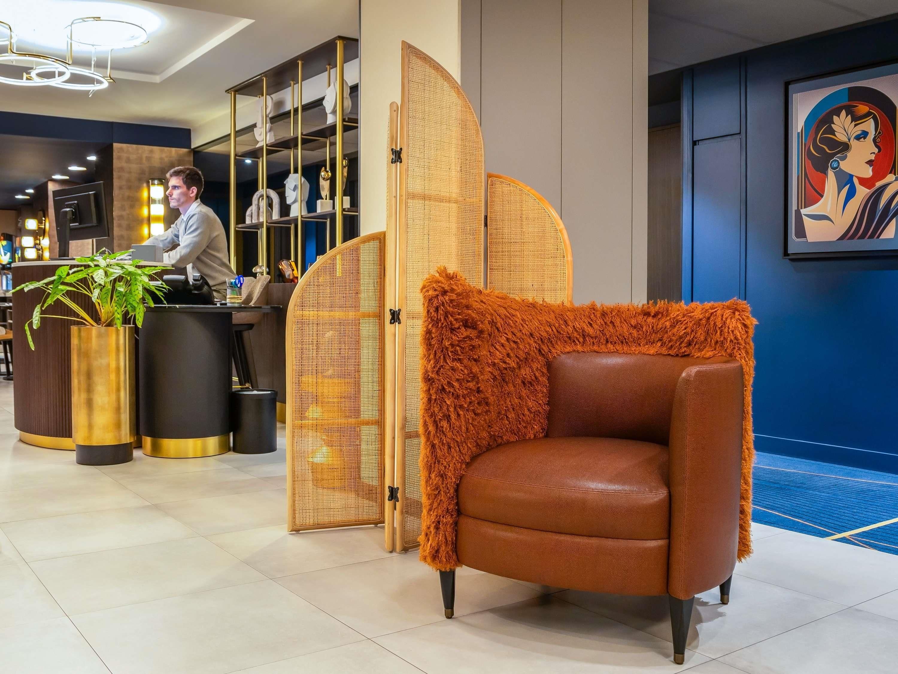 Un área de recepción con mostrador, una zona de estar con sillón de cuero marrón, una planta decorativa y un cuadro grande en la pared.