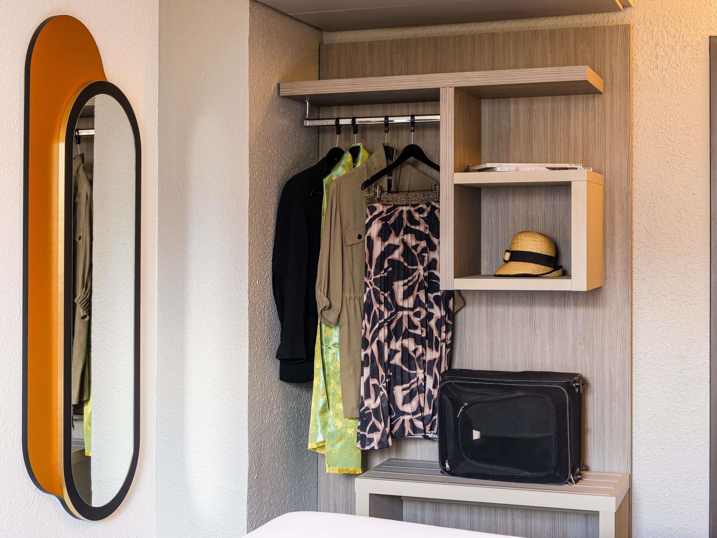 Habitación de hotel con armario, espejo, perchero con ropa y estante con sombrero y maleta.