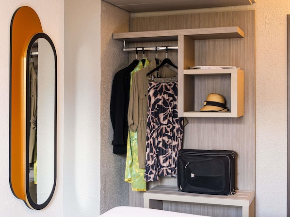 A hotel room with a wardrobe, a mirror, a hanging rack with clothes, a shelf with a hat, and a suitcase. - ibis Styles Saint-Quentin (Saint-Quentin)