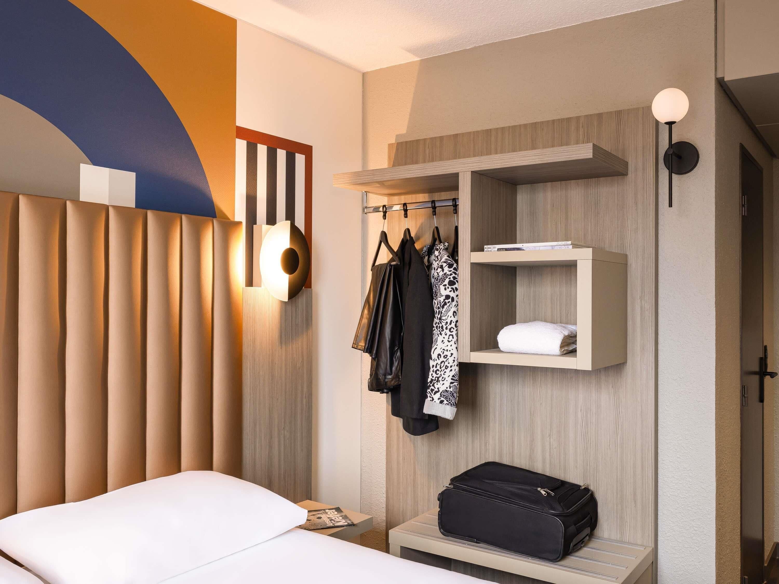 A hotel room with a bed, a bedside table with a magazine, a hanging coat rack, a shelf with a towel, and a suitcase.