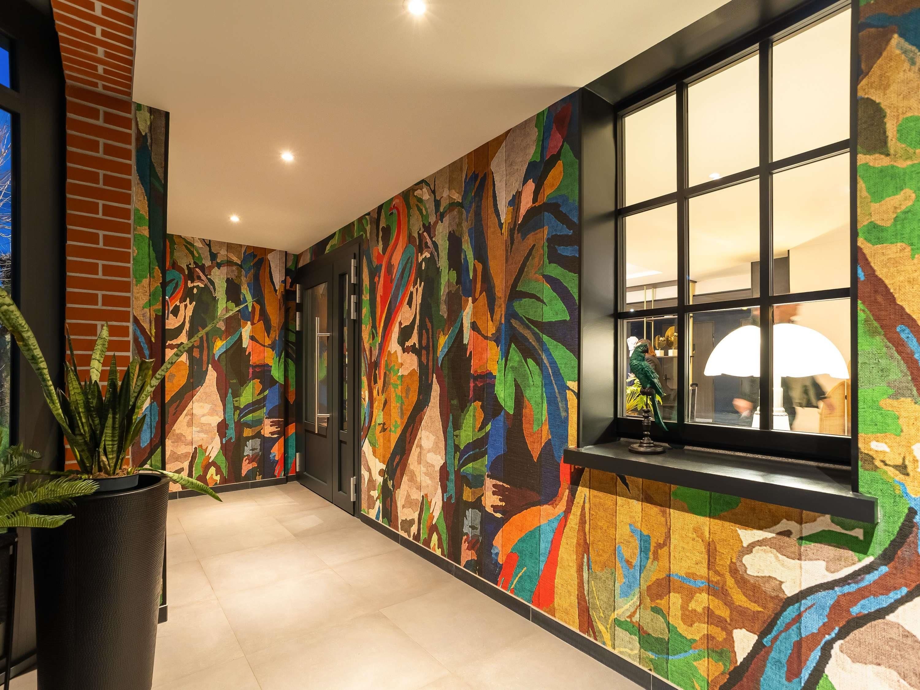 A hallway with a colorful abstract mural on the walls, a potted plant, and a window with a view of the interior.