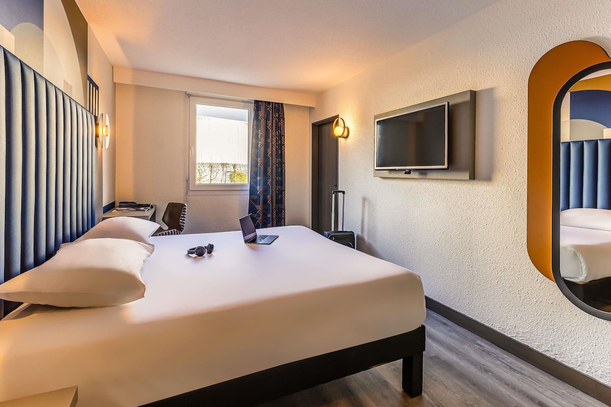 A hotel room with a bed, a TV mounted on the wall, a window with curtains, and a small desk with a laptop and headphones.