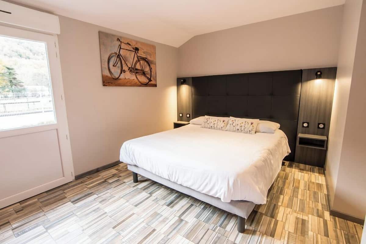 A bedroom with a large bed, a headboard with integrated lighting, a bicycle artwork on the wall, and a window with a view of greenery.